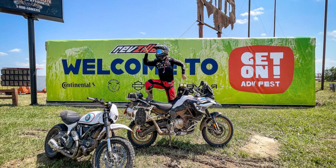 Rider posing on dirt bike in front of banner for GET ON Adv Fest by RevZilla