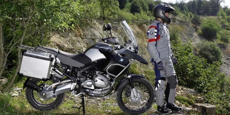 A Beemer dismounted from his BMW and standing in front of a wall of green foliage. Media sourced from Beach Moto.