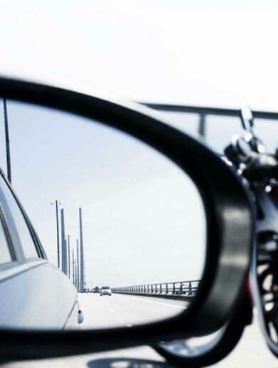 A car mirror with a motorcyclist int he background. Media sourced from RideApart.