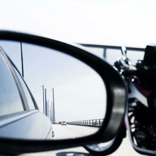 A car mirror with a motorcyclist int he background. Media sourced from RideApart.