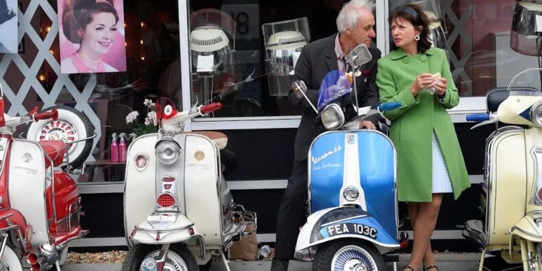 An old Italian couple next to a lineup of classic Vespa scoots. Media sourced from The Guardian.